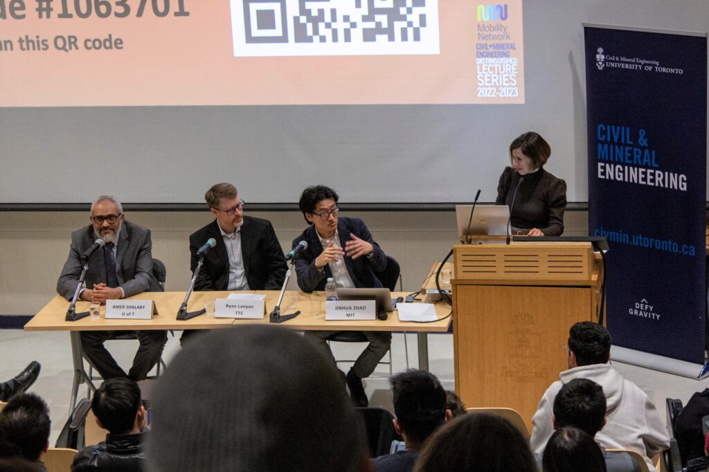 Three panelists sit at table, moderator at podium. View from the back of the hall.